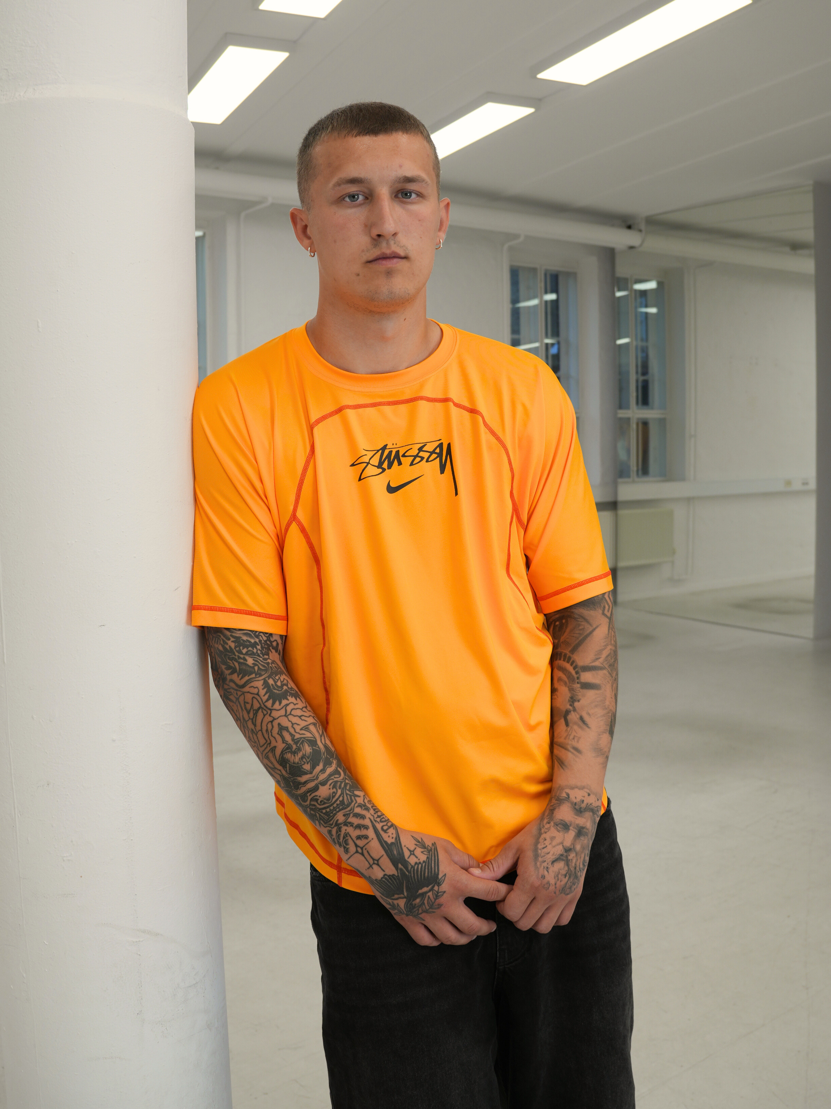 Man i orange t-shirt med tatueringar lutar sig mot vit pelare