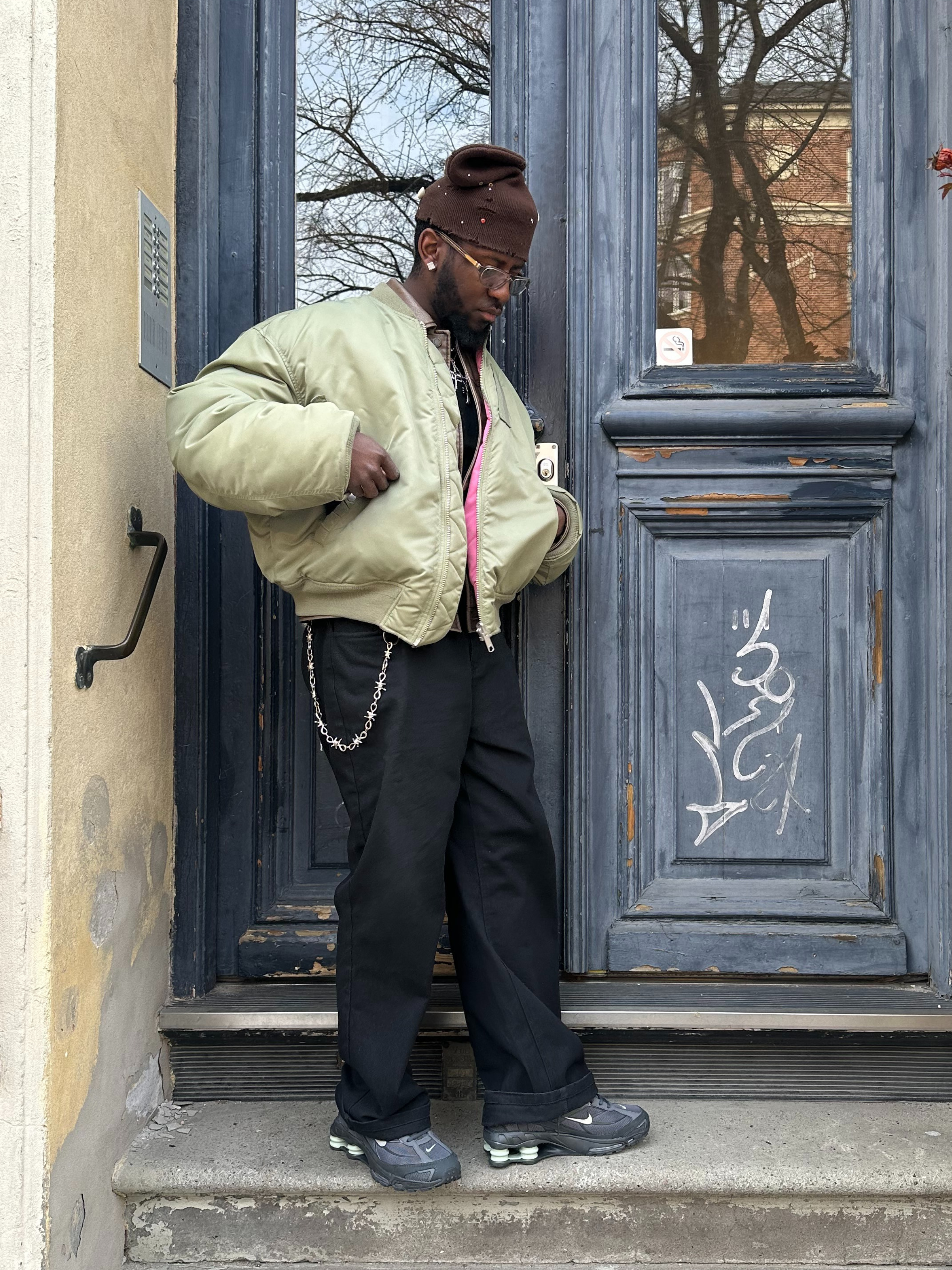 Man i olivgrön jacka, svarta byxor och grå skor poserar framför dörr.